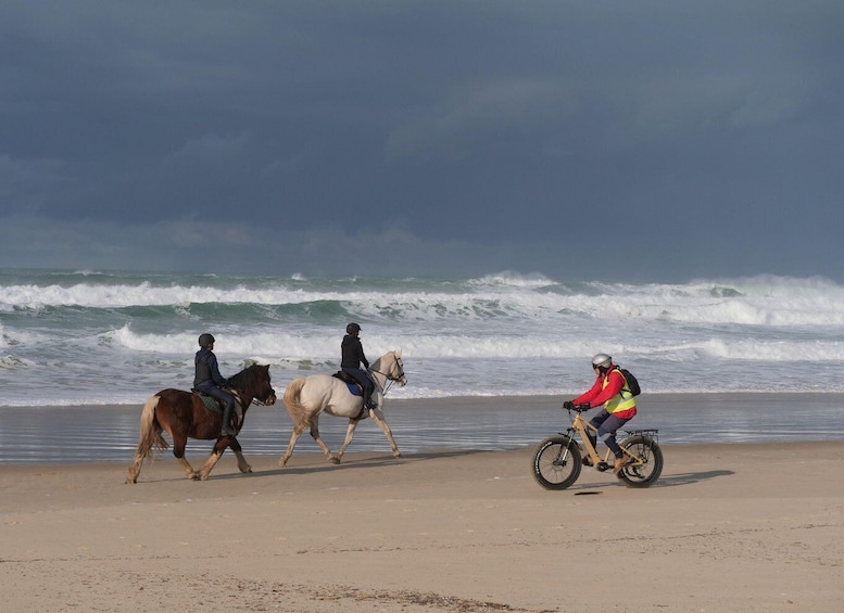 Three people riding horses and one person on a bicycle on a beach with waves in the background. 5