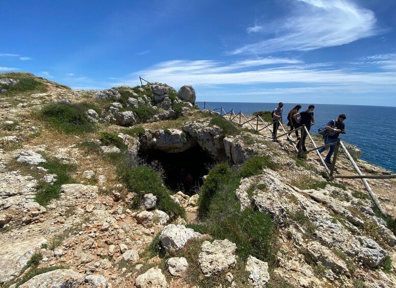 A rocky coastal landscape with a cave entrance, a wooden fence, and a group of hikers. 2
