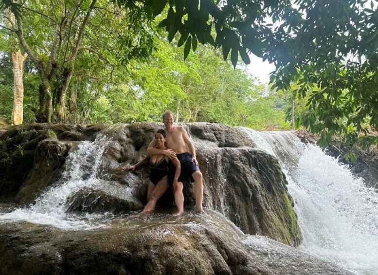 Two people standing on a rock by a waterfall surrounded by greenery. 3