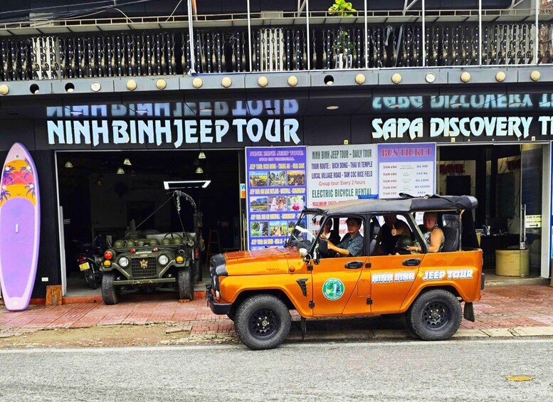 A jeep with passengers, parked in front of a building with signs for a jeep tour and a surfboard. 1