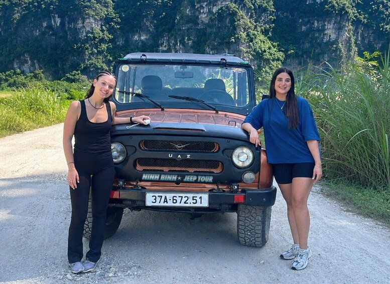 Two people standing beside a UAZ vehicle with a license plate reading '37A-672.51' on a dirt road with greenery and cliffs in the background. 2
