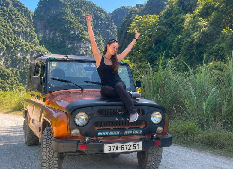 A person sitting on a Jeep with raised arms, surrounded by lush greenery and mountains. 5