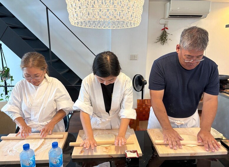 Three people rolling dough on wooden boards in a kitchen with a hanging light fixture. 3