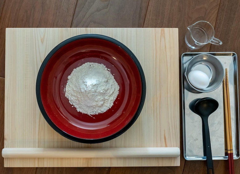 A red bowl with flour, a measuring cup, a metal bowl with a white substance, and chopsticks on a wooden board. 2