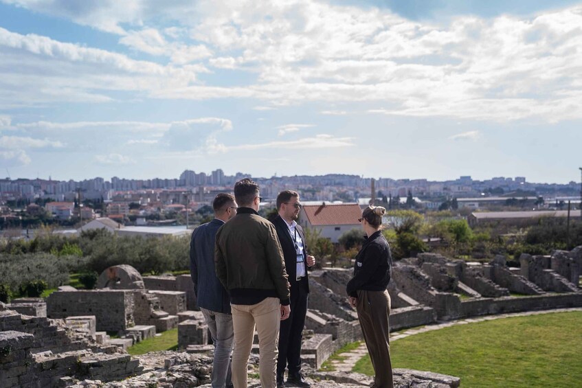 A group of people standing at an archaeological site with ruins and a cityscape in the background. 5