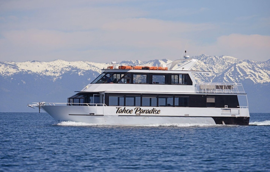 A white boat named Tahoe Paradise on a body of water with snow-capped mountains in the background. 1
