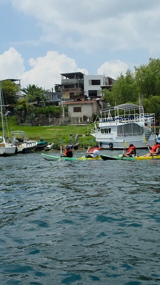 A calm waterway with several boats, including kayaks and a sailboat, docked near a residential area with houses and greenery. 1
