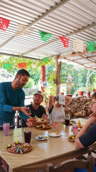 A group of people dining outdoors under a metal roof, with tables set with food and drinks, surrounded by greenery and decorative paper lanterns. 4