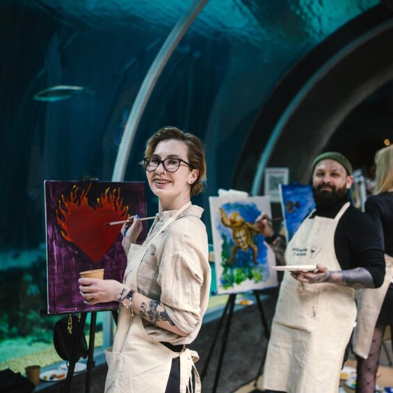 A person holding a painting of a heart with flames, standing in an indoor art gallery with other artworks on display. 1