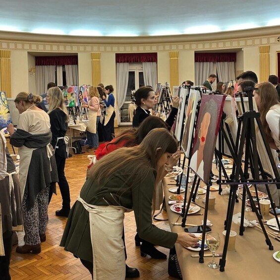 A group of people engaged in painting on easels in a room with wooden floors and red curtains. 3