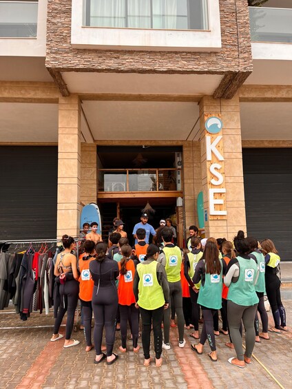 A group of people in wetsuits standing in front of a building with a sign that reads 'KITE & SURF'. 3