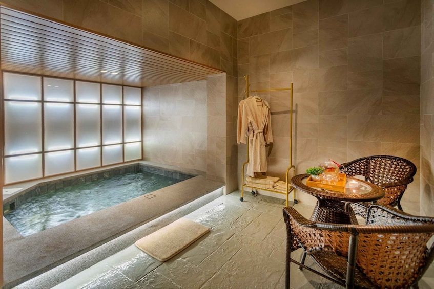 A spa room with a hot tub, a wooden bath mat, wicker chairs, and a towel rack. 4
