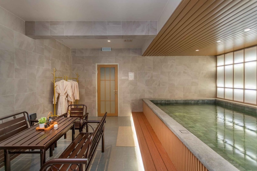 A spa room with a wooden bench, a small table with a plant, a towel rack, and a large indoor pool. 5