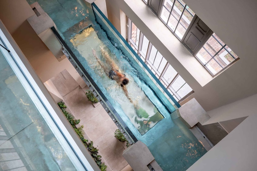 A swimming pool with a person swimming, surrounded by indoor architecture and large windows. 1