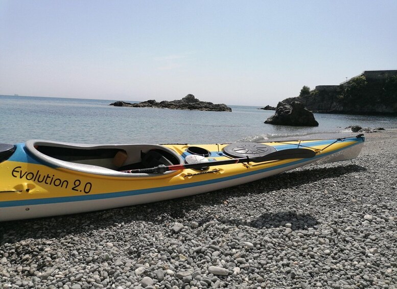 A yellow kayak with 'Evolution 2.0' written on it, resting on a pebble beach with calm water and rocky outcrops in the background. 3