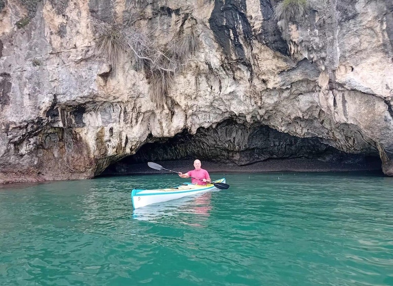 A person kayaking in a turquoise kayak with a paddle, surrounded by a rocky cave entrance and calm water. 1