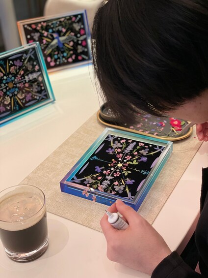 A person examining a framed floral artwork on a table, with a glass of dark liquid beside them. 4