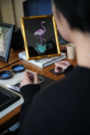 A person using a smartphone at a desk with a framed artwork of a flamingo, a cup, and small containers. 2