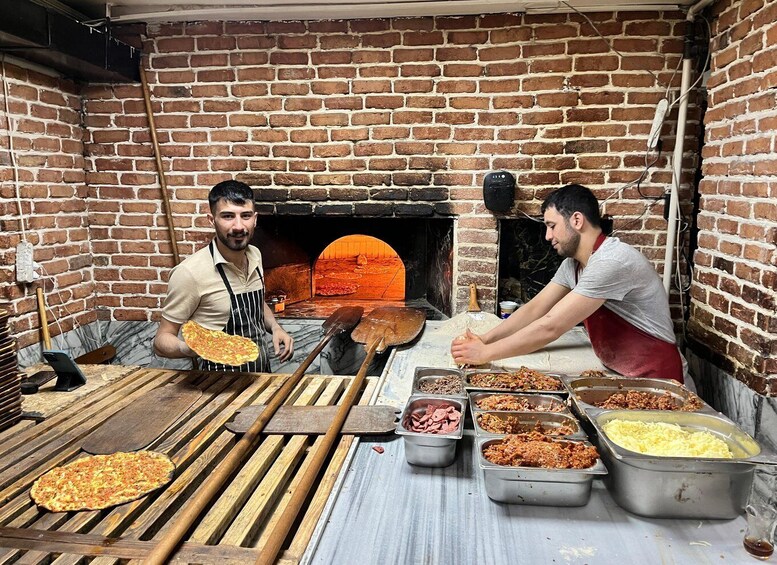 Two people preparing pizzas in a brick oven kitchen with various toppings and tools. 3