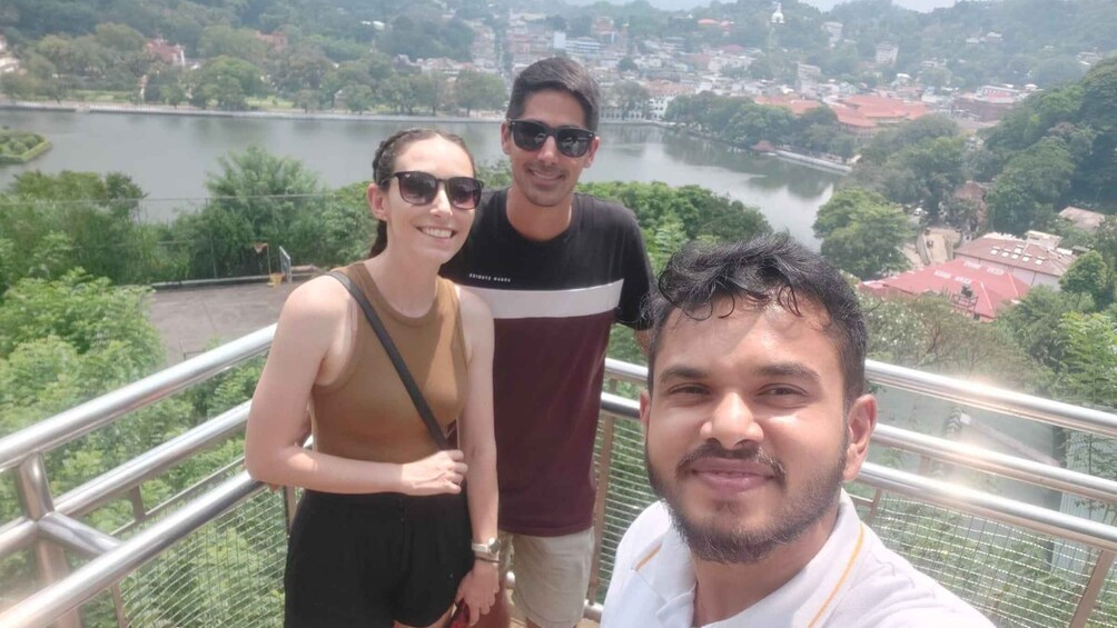 Three people standing on a balcony with a river and buildings in the background. 1