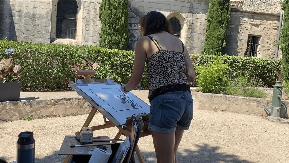 A person painting on an easel in an outdoor setting with stone buildings and greenery in the background. 3