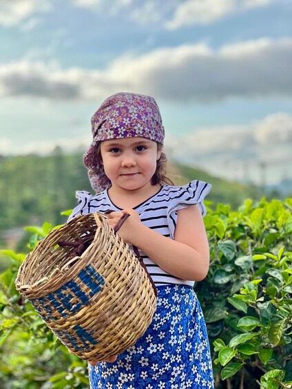 A child holding a woven basket, wearing a floral dress and a patterned headscarf, standing in front of a green hedge. 3