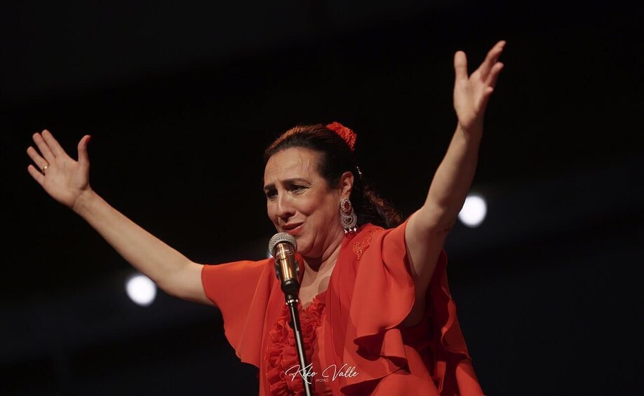 A person in a red dress with ruffled sleeves, standing with arms raised, and a microphone in front. 1
