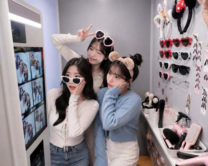 Three people posing in a room with a mirror, a wall with heart-shaped items, and a shelf with hair care products. 4