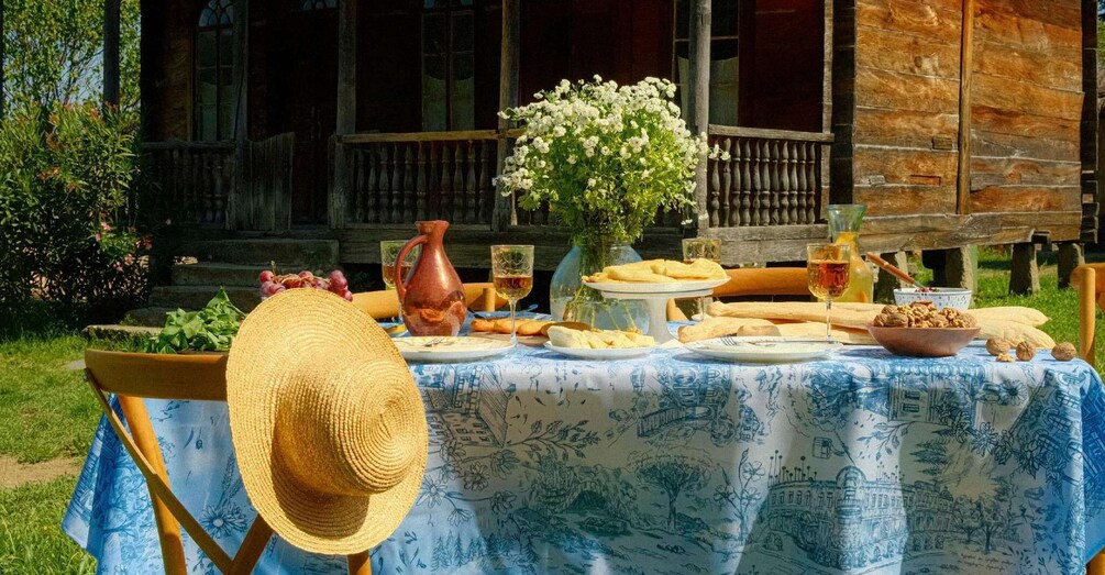 An outdoor picnic with a table set for two, featuring a variety of foods and drinks, a vase of flowers, and a rustic wooden building in the background. 3