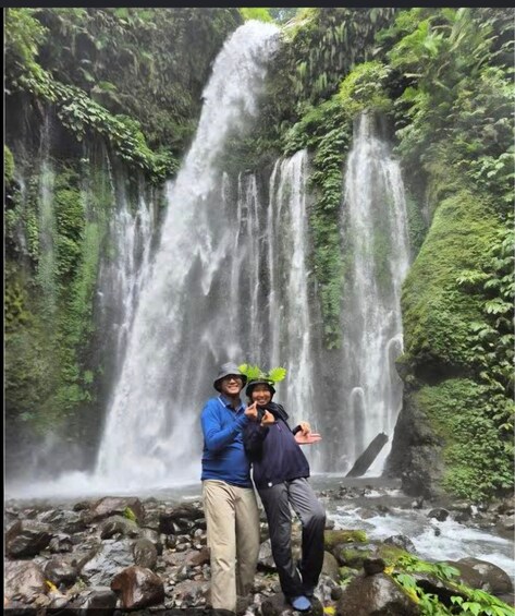 Two people posing in front of a waterfall with lush greenery. 1