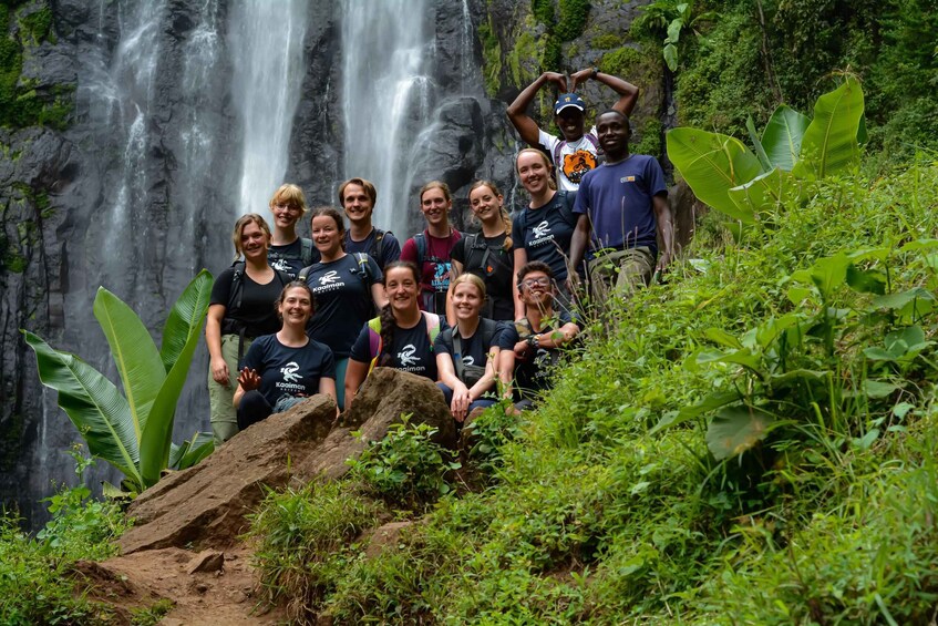 A group of people posing in front of a waterfall with lush greenery. 1