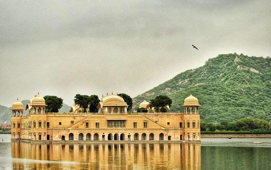 A historic building with domed architecture, situated on a reflective water body, with a forested hill in the background. 2