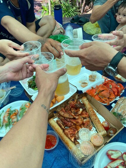 A group of people toasting with plastic cups, a variety of food including seafood and grilled items, and a blue tablecloth. 3