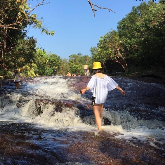 A person in a white blouse and yellow hat walking through a shallow, rocky stream surrounded by trees. 1