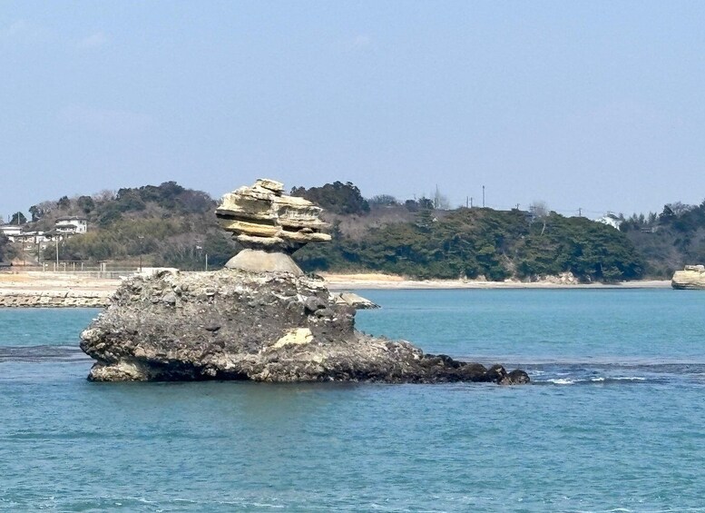 A rock formation balanced on a smaller rock in the sea, with a beach and buildings in the background. 2