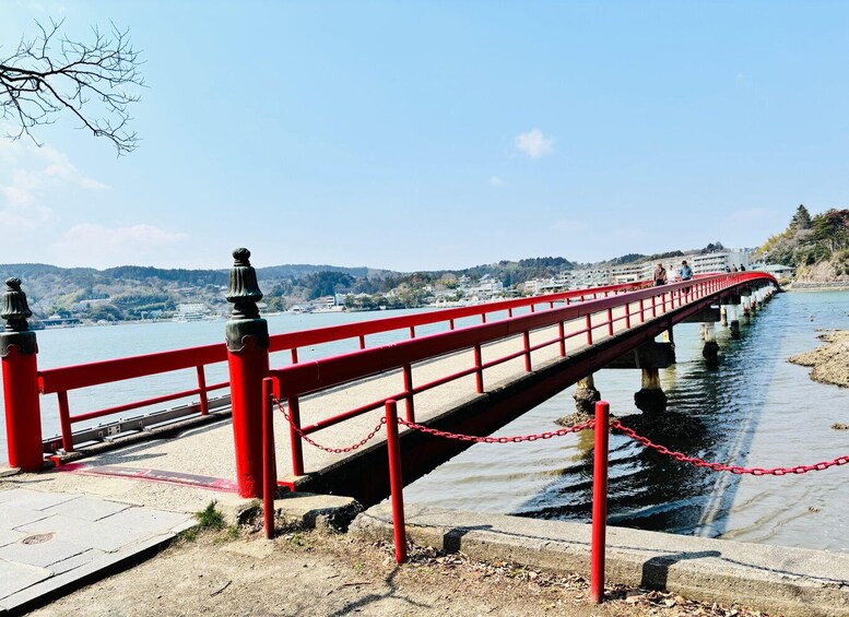 A red bridge over water with a chain barrier, a clear sky, and distant buildings. 1