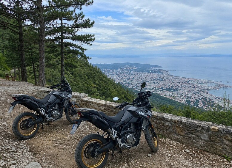 Two motorcycles parked on a rocky hillside overlooking a coastal cityscape. 1