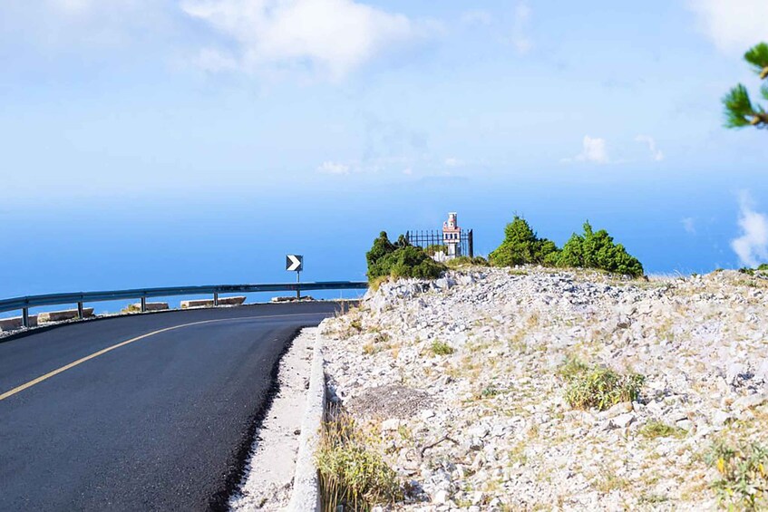 A coastal road curving to the right with a guardrail, rocky terrain, and a lighthouse in the distance. 5