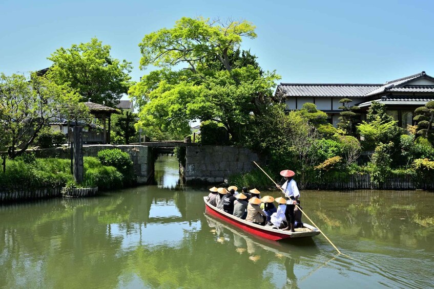 A traditional boat with passengers wearing conical hats, navigating a calm river with bridges and buildings in the background. 2