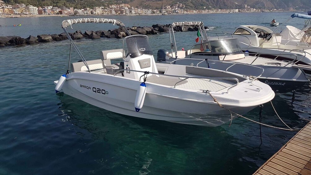 A white motorboat named BARQ A Q20 docked at a marina with a rocky breakwater and a coastal town in the background. 1