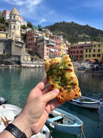 A hand holding a piece of food with a scenic waterfront and colorful buildings in the background. 1