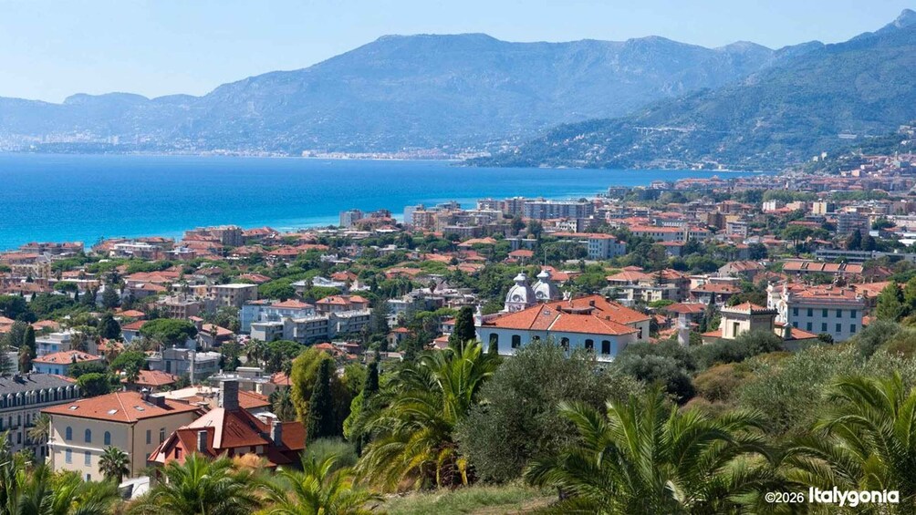 A coastal cityscape with red-roofed buildings, a clear blue sea, and mountains in the background. 5