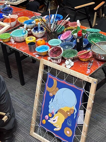 A table with various paint colors and brushes, and a framed artwork of a blue elephant. 3