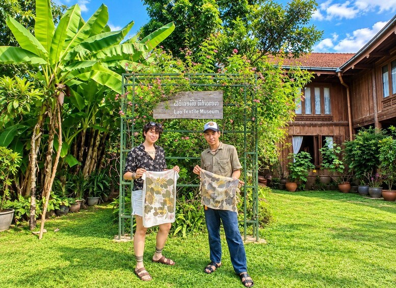 Two people holding a fabric with a floral pattern in front of a Lao Textile Museum sign. 3