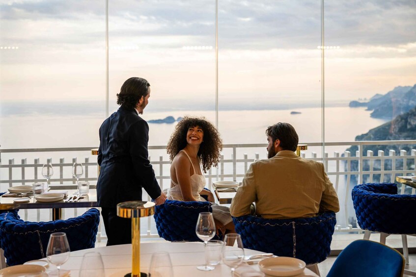 A restaurant with a view of the sea, blue upholstered chairs, and a waiter serving. 2