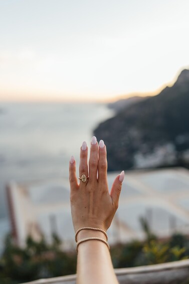 A hand with a ring and bracelet, overlooking a coastal landscape with mountains and a beach. 3