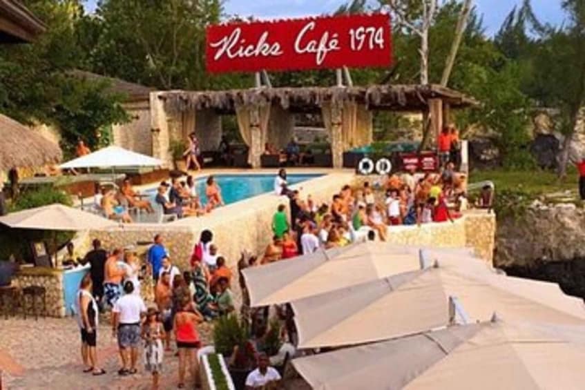 A crowded outdoor swimming pool area with a sign reading 'Ricks Café 1974' and people relaxing around the pool. 3