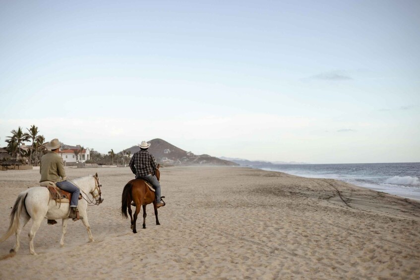 Two people riding horses on a beach with ocean waves and a mountain in the background. 1