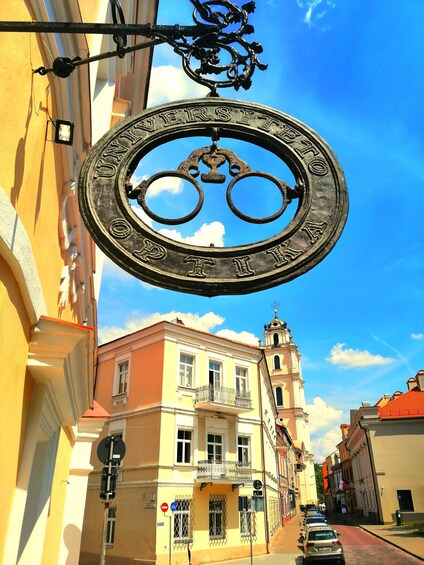 A street sign with 'UNIVERSITY OF ATHENS' inscribed on it, a building with a red roof, and a clear blue sky. 1