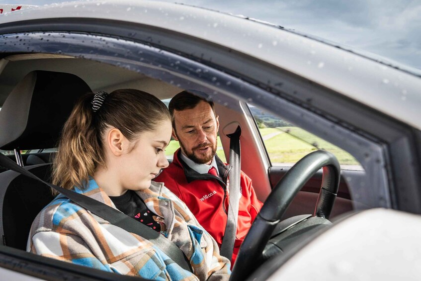A person in a red jacket sitting in a car, with another person in a plaid shirt in the driver's seat. 4